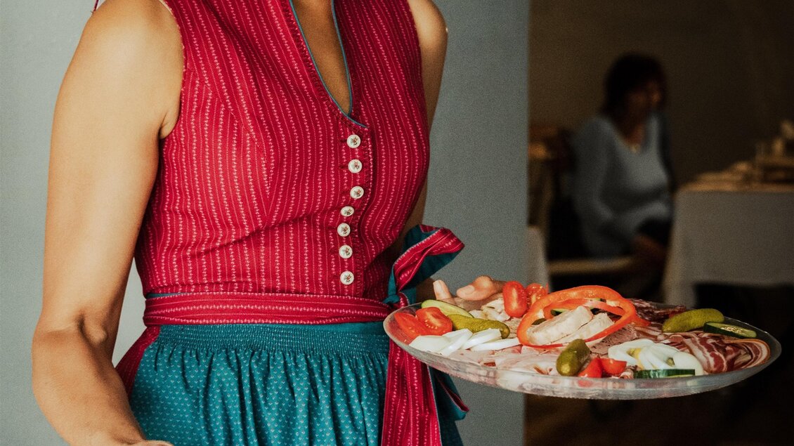 Eine lächelnde Frau trägt zwei große Teller mit frisch zubereiteten Speisen. Sie trägt ein knalliges Outfit mit rosa und türkisfarbenen Akzenten. | © Buschenschank Hildebrand