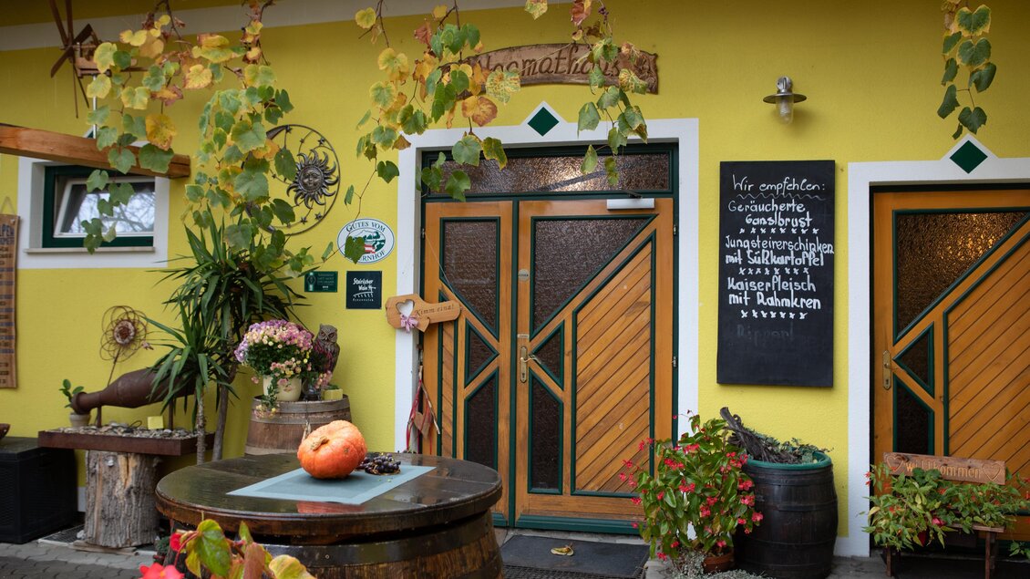Ein einladendes, gelbes Gebäude mit Holzverzierungen und einer Eingangstür. Vor der Tür stehen Pflanzen und eine Tafel mit Schrift. | © GlaMUR-Genuss am Fluss