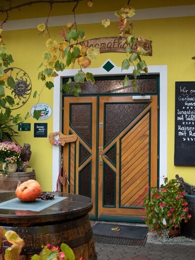 An inviting yellow building with wooden decorations and a front door. Plants stand in front of the door along with a sign with writing. | © GlaMUR-Genuss am Fluss