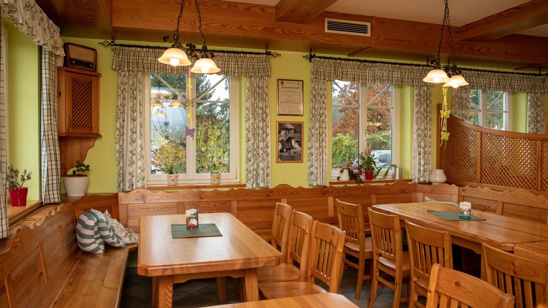 Ein gemütlicher Restaurantbereich mit Holztischen und Bänken. Die Wände sind in einem warmen Gelbton gestrichen und große Fenster lassen viel Tageslicht herein. | © GlaMUR-Genuss am Fluss