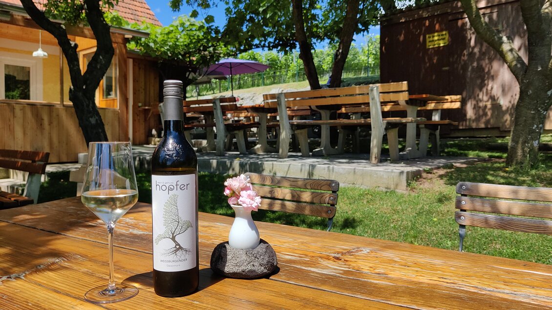 Ein Holz Tisch im Freien mit einer Flasche Wein und einem Glas. Im Hintergrund sind Sitzgelegenheiten und Bäume zu sehen.