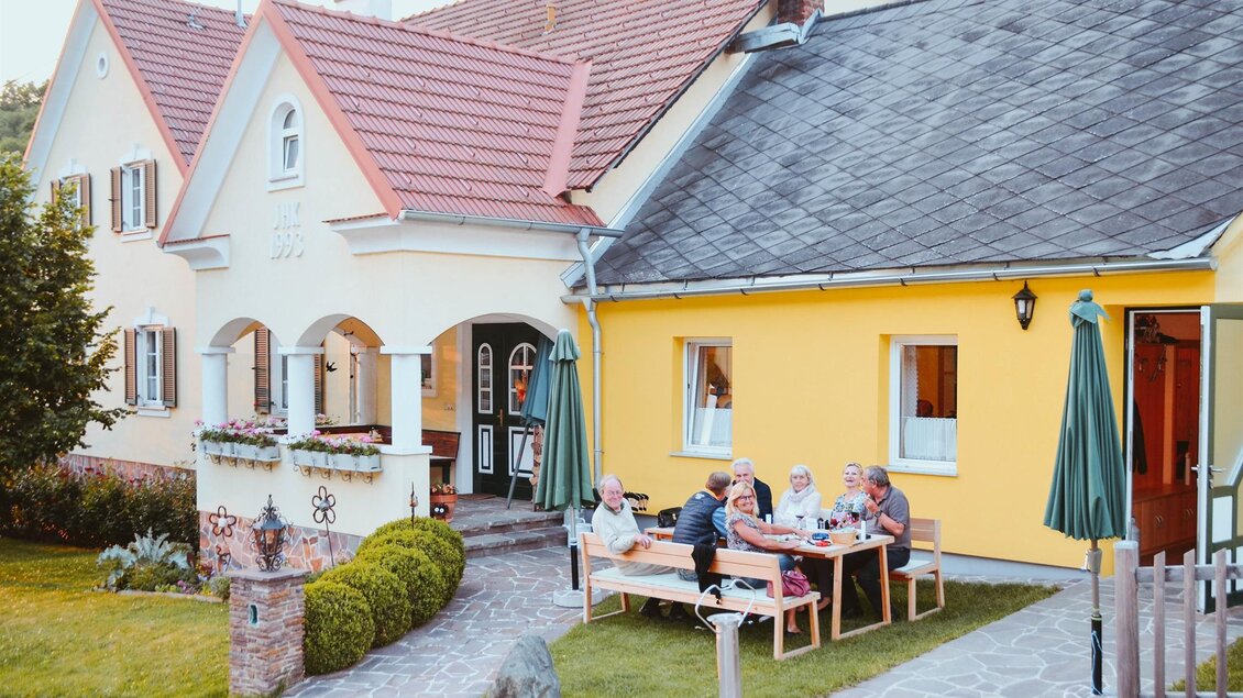Ein einladendes Haus mit gelber Fassade und einem Garten. Eine Gruppe von Menschen sitzt draußen an einem Tisch und genießt die Zeit zusammen. | © Lederer