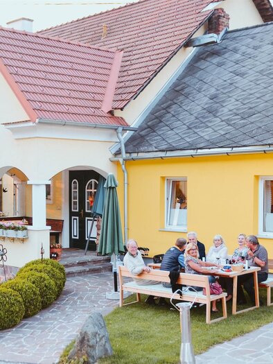 An inviting house with a yellow facade and a garden. A group of people is sitting outside at a table and enjoying their time together. | © Lederer
