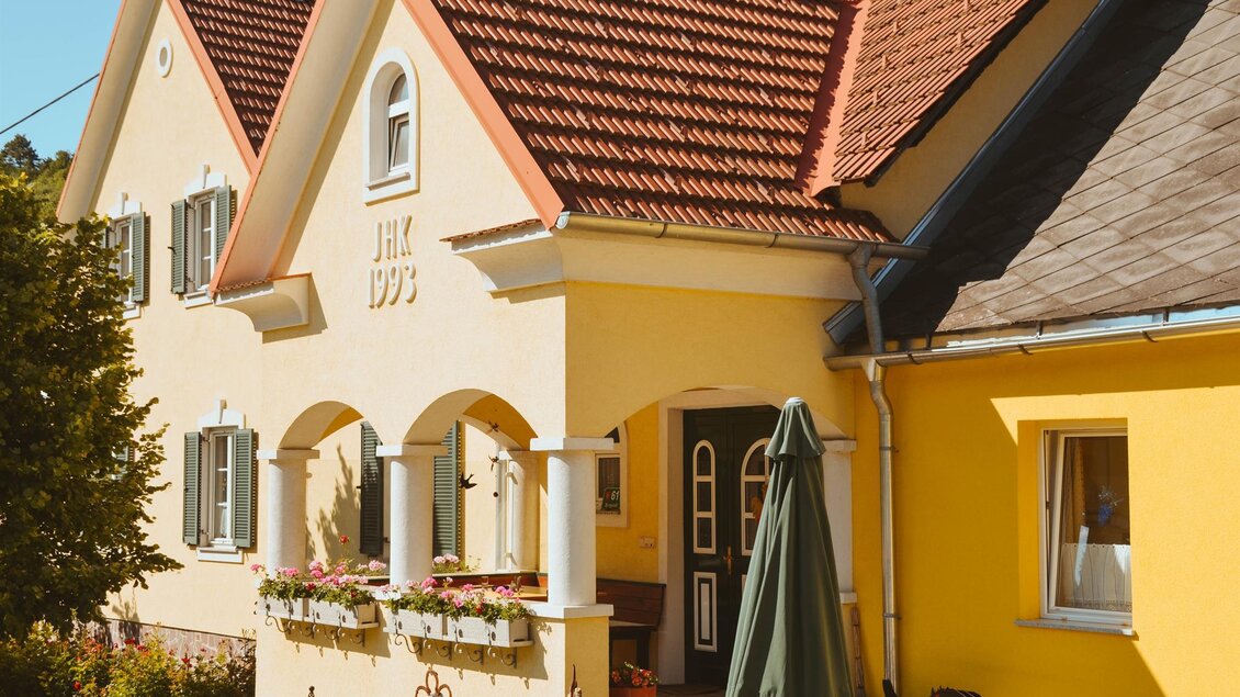 Ein charmantes, gelbes Haus mit einem roten Dach und weißen Säulen. Die Fenster sind mit grünen Fensterläden versehen und es gibt einen Schirm vor der Eingangstür. | © Lederer
