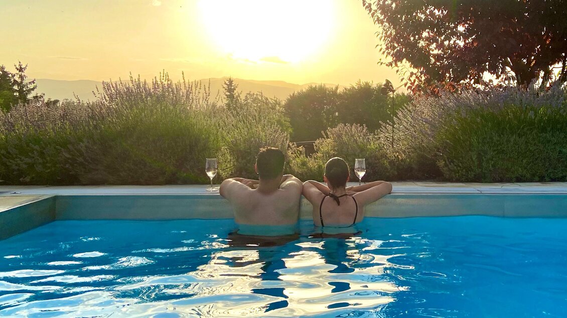 Ein Paar sitzt entspannt am Rand eines Pools und genießt den Sonnenuntergang. Beide halten ein Glas in der Hand und schauen in die Weite. | © Brigitte Maier | Klapotetzhof Silberschneider