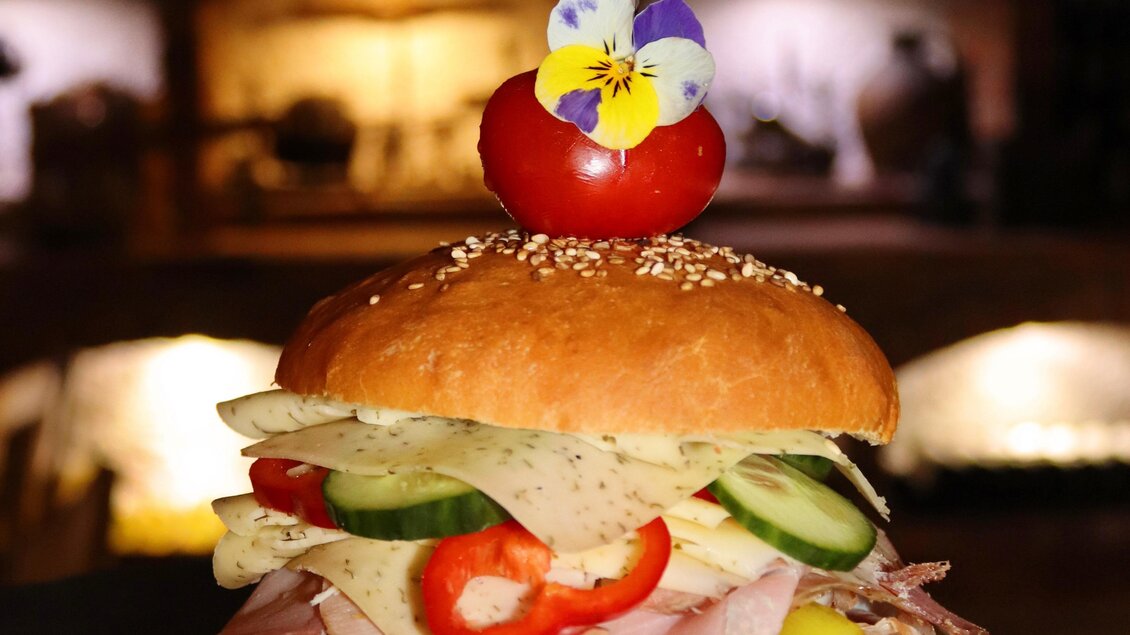 Ein appetitlicher Hamburger mit Schinken, Käse, Gurken und Paprika. Oben ist eine Kirschtomate und eine essbare Blüte dekorativ platziert. | © Klapotetzhof Silberschneider