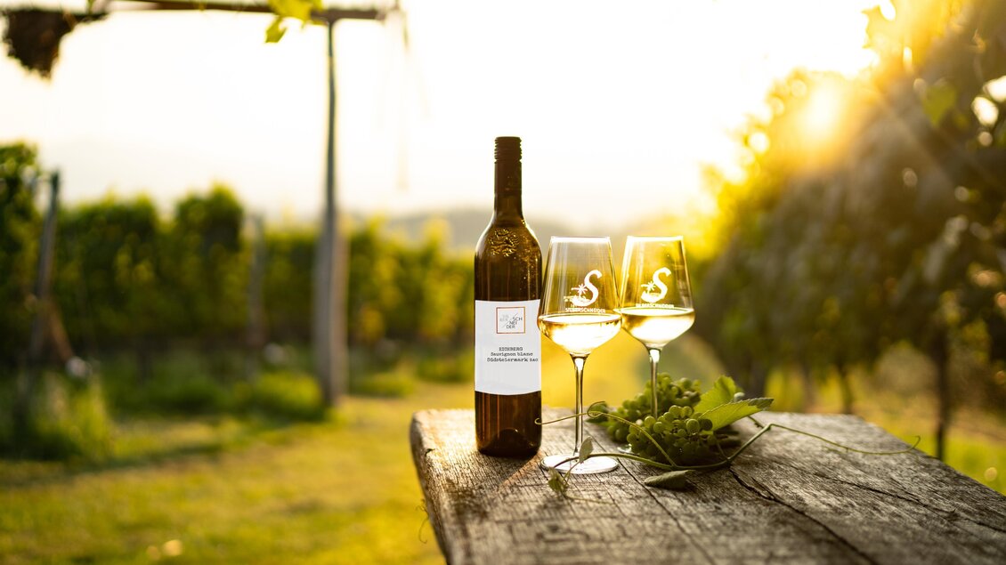 Eine Flasche Weißwein steht auf einem Holztisch neben zwei Gläsern. Im Hintergrund sind Weinreben und die Sonne strahlt warm auf die Szene. | © Alois Stingl | Klapotetzhof Silberschneider