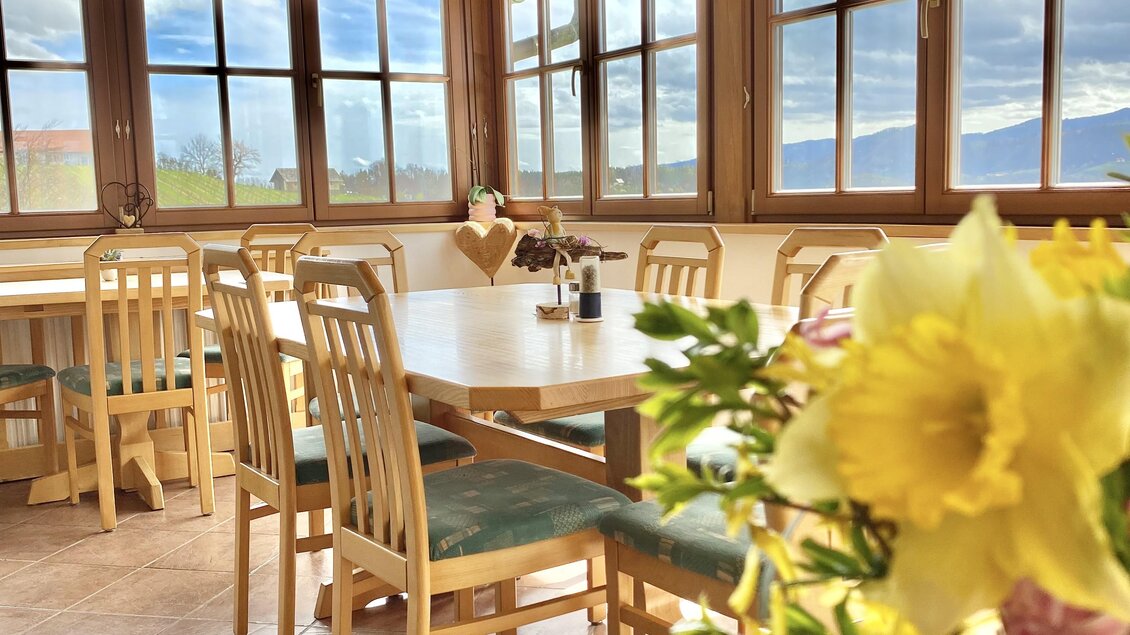 Ein einladendes Restaurant mit hellen Fenstern und holzverkleideter Decke. Auf dem Tisch steht eine Blumenvase, die eine freundliche Atmosphäre schafft.