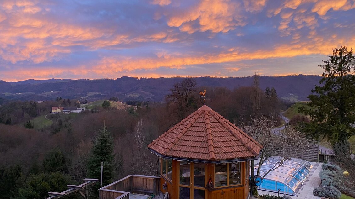 Eine panoramaartige Aussicht mit einem Holzanbau und einem Schwimmbad. Der Himmel ist in warmen Farben während des Sonnenuntergangs gefärbt.