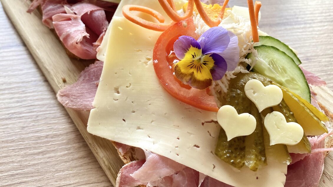 Ein schön dekoriertes belegtes Brot mit Schinken, Käse, Gurken und frischen Blumen. Die Anordnung ist appetitlich und farbenfroh.