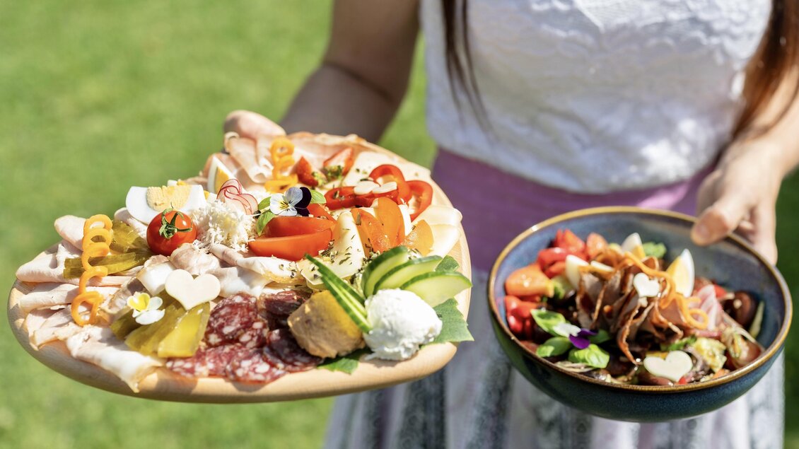 Eine Person hält zwei Platten mit Essen. Die erste Platte enthält eine bunte Auswahl an Wurst, Käse und Gemüse, während die zweite eine frische Salatmischung zeigt. | © © RMSW GmbH/ Nikola Milatovic