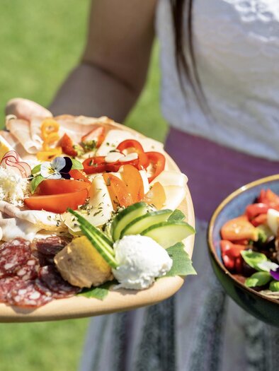 A person holds two plates with food. The first plate contains a colorful selection of sausage, cheese, and vegetables, while the second shows a fresh salad mix. | © © RMSW GmbH/ Nikola Milatovic