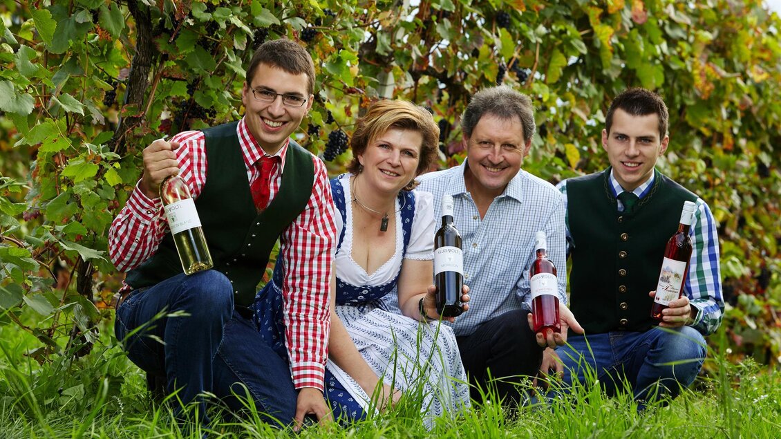 Eine Familie steht in einem Weinfeld und hält Flaschen Wein in der Hand. Sie tragen traditionelle Kleidung und lächeln in die Kamera. | © Foto Melbinger Klug-Voltl