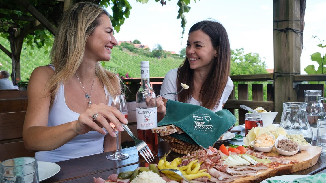 Zwei Frauen sitzen an einem Tisch im Freien und genießen ein reichhaltiges Essen. Sie lächeln und halten Gläser mit einem Roséwein in der Hand. | © Schilcherland Steiermark