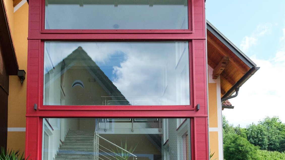 Ein modernes Haus mit einer auffälligen roten Fassade und großen Fenstern. Der Eingang ist freundlich gestaltet und von Pflanzen umgeben. | © Schilcherland Steiermark