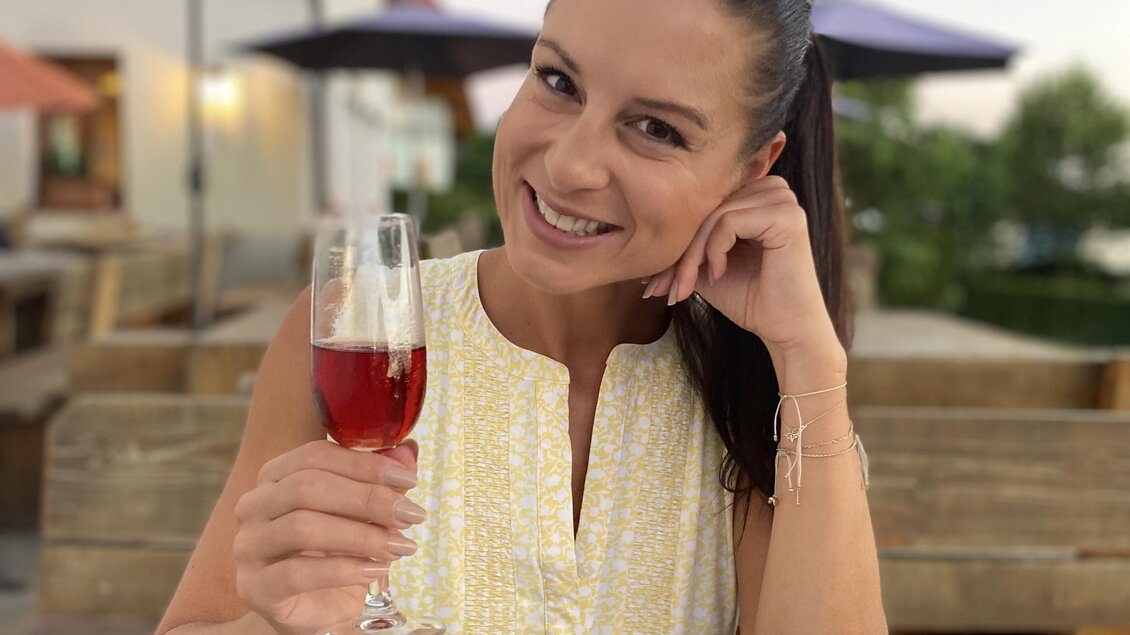 Eine lächelnde Frau mit einem Glas Getränk sitzt an einem Holztisch im Freien. Im Hintergrund sind Sonnenschirme und eine entspannte Atmosphäre zu sehen. | © silviaschreibt.de