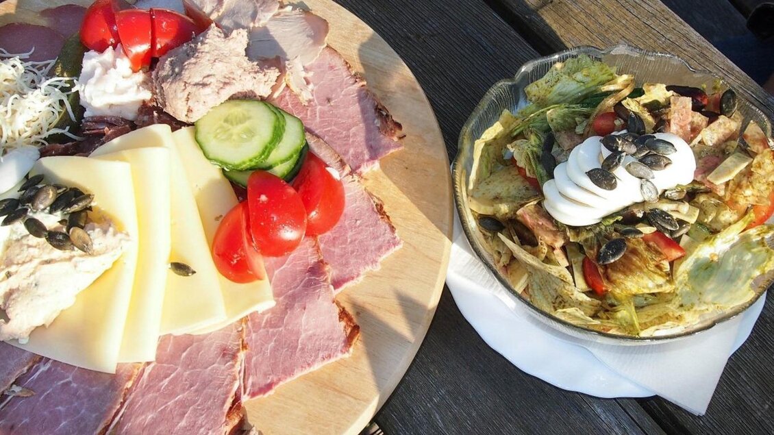 Ein Teller mit Wurst, Käse, Tomaten und Gurken, begleitet von einem frischen Salat mit Joghurtdressing und Kürbiskernen. Das Gericht ist appetitlich angerichtet und erinnert an ein herzhaftes Mittagessen. | © Weingut Koller
