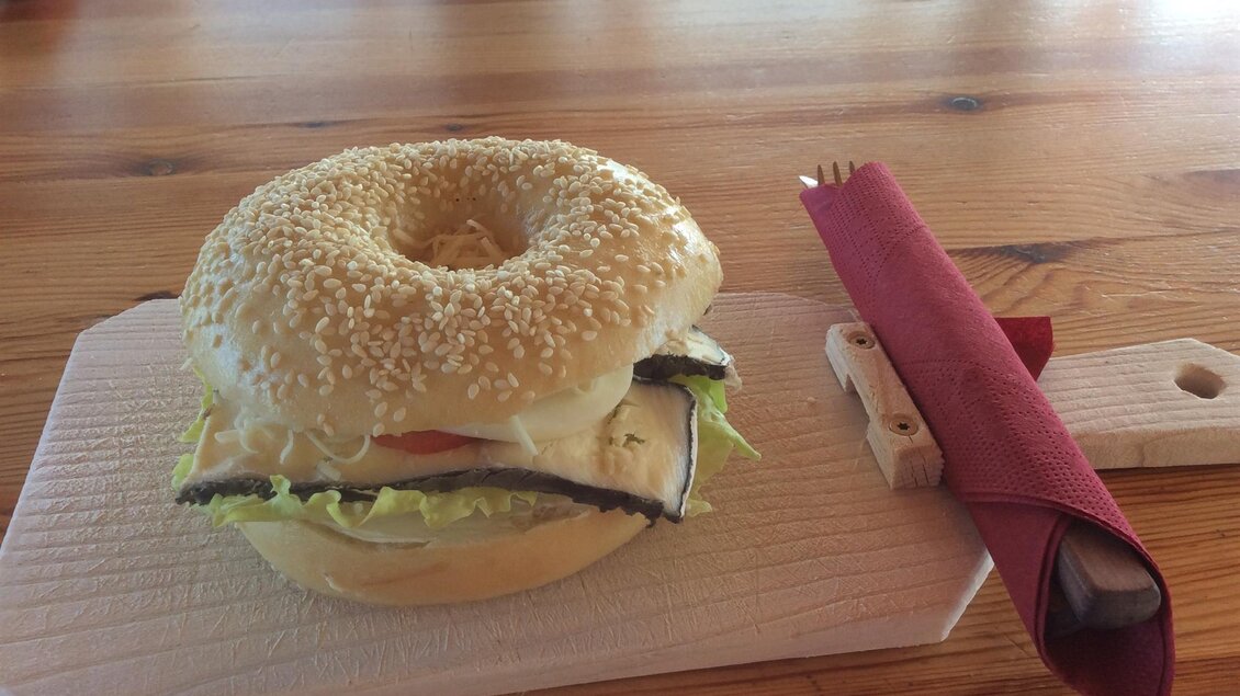 Ein Bagel mit Sesam, gefüllt mit frischem Salat, Tomaten und einer weißen Creme. Daneben liegt ein Besteckset auf einem Holzbrett. | © Weingut Koller