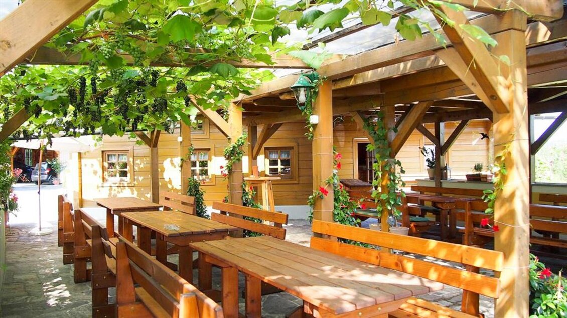 Eine gemütliche Gaststätte mit Holzterrasse und Platz für Gäste. Grünpflanzen wachsen über den Tischen und schaffen eine einladende Atmosphäre. | © Marion & Peter Labanz | Buschenschank Labanz