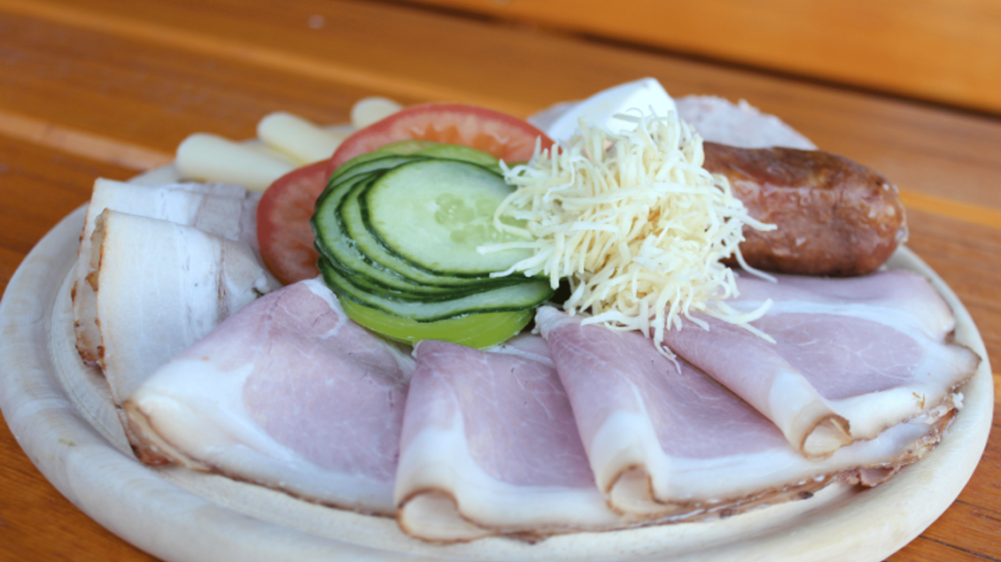 Ein Teller mit verschiedenen Wurst- und Käsesorten, garnierte mit Gurken, Tomaten und etwas Sauerrahm. Die Präsentation ist ansprechend auf einem Holzbrett angerichtet. | © Marion & Peter Labanz | Buschenschank Labanz