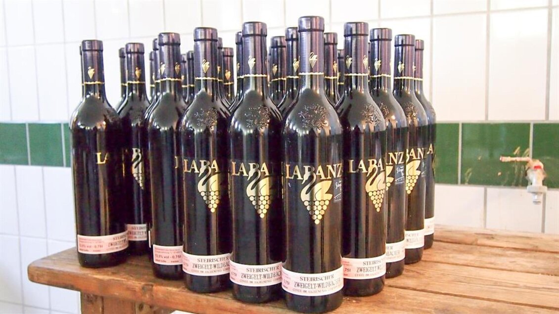 Eine Reihe von Weinflaschen steht ordentlich auf einem Holzregal. Die Flaschen sind dunkel mit einem auffälligen Etikett. | © Marion & Peter Labanz | Buschenschank Labanz