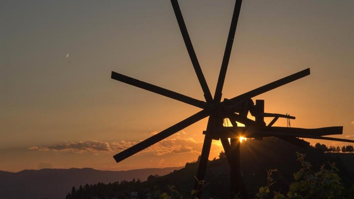 Ein Windrad silhouettiert vor einem Sonnenuntergang. Die warmen Farben des Himmels spiegeln sich im Hintergrund wider. | © Weingut Lambauer