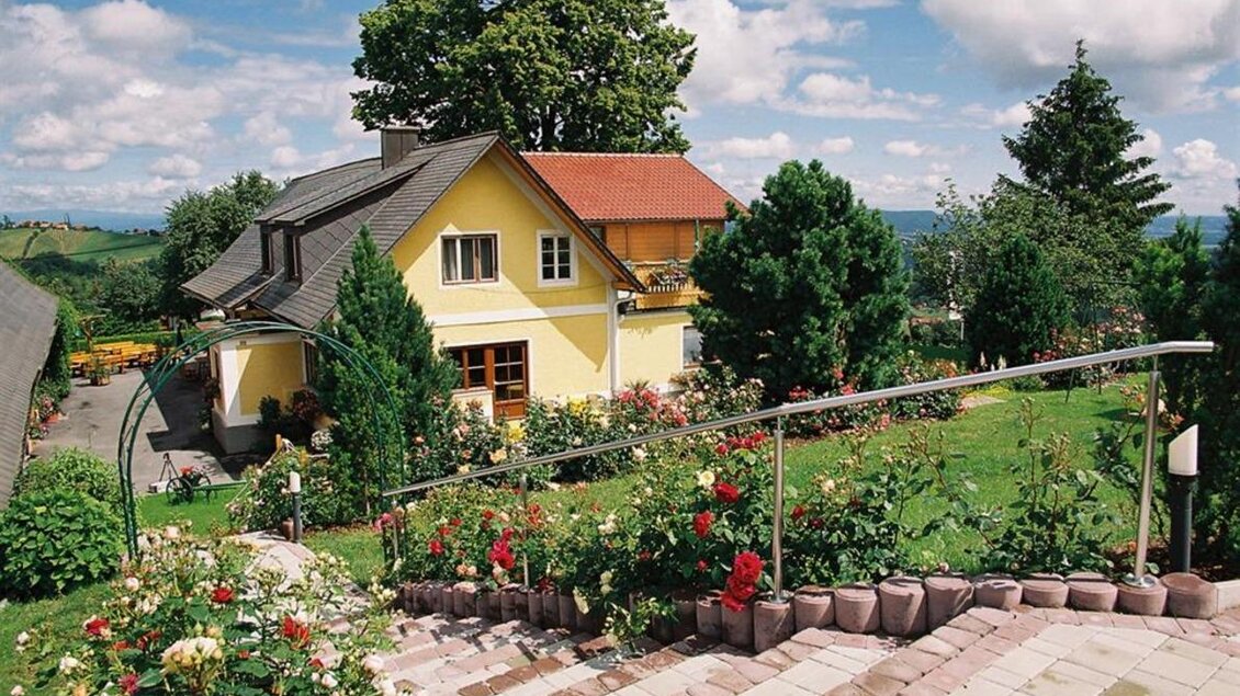 Ein schönes gelbes Haus umgeben von Gartenpflanzen und Blumen. Der Himmel ist klar und die Landschaft ist grün und einladend. | © Weingut Lambauer