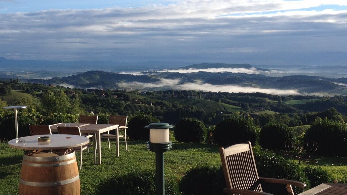 Eine wunderschöne Aussicht auf die hügelige Landschaft mit Nebel im Tal. Im Vordergrund stehen Gartenmöbel und ein Weinfass. | © Weingut Lambauer