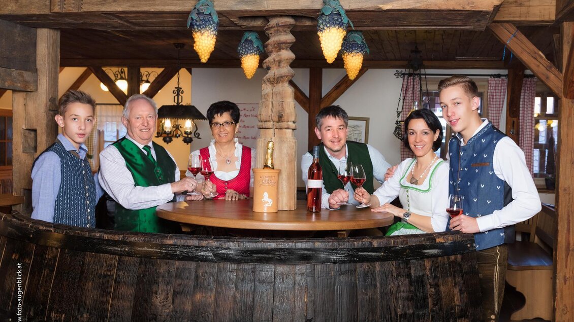 Eine Gruppe von sechs Personen steht an einer rustikalen Bar. Sie tragen traditionelle Trachten und halten Gläser in der Hand, lächelnd und in geselliger Stimmung. | © Weingut & Buschenschank Lazarus
