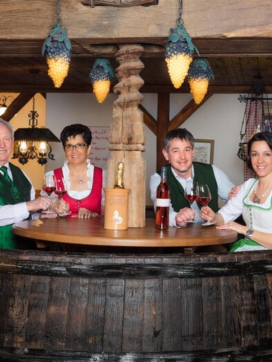 A group of six people is standing at a rustic bar. They are wearing traditional costumes and holding glasses in their hands, smiling and in a festive mood. | © Weingut & Buschenschank Lazarus