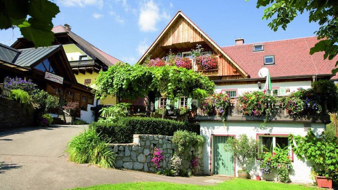Ein malerisches Bauernhaus mit blühenden Balkonen und viel Grün. Der Himmel ist klar und die Umgebung ist ruhig und einladend. | © Weingut & Buschenschank Lazarus