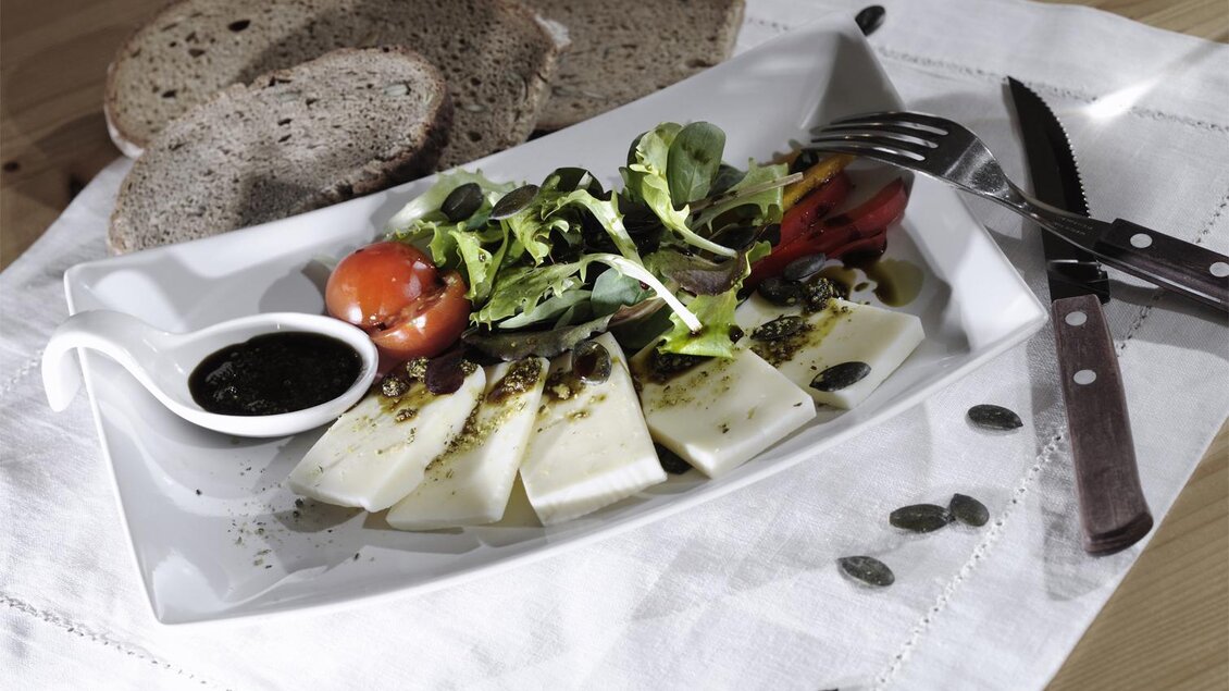 Ein Teller mit frischem Käse, gemischtem Salat und einer kleinen Schale mit Dressing. Im Hintergrund liegen zwei Scheiben Brot auf einem Tisch. | © www.michilorenz.at