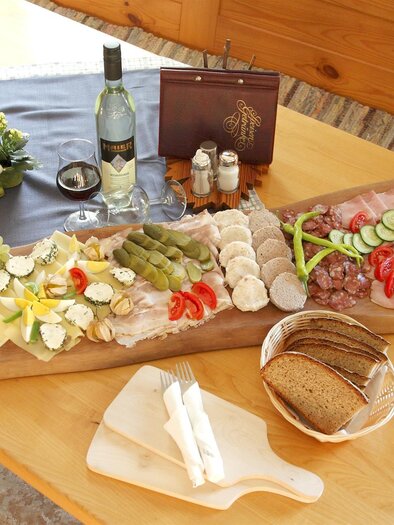 A rich platter with sausage, cheese, grapes, and vegetables is on a wooden table. Next to it are bread and a bottle of wine. | © Weingut Maier
