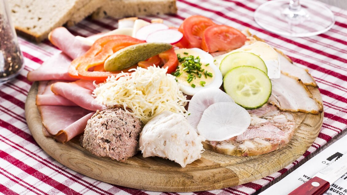Ein Holzbrett mit einer Auswahl an Wurst, Käse, frischem Gemüse und Brot. Dazu ein Glas Wein und ein kariertes Tischdeckchen. | © Weingut Malli