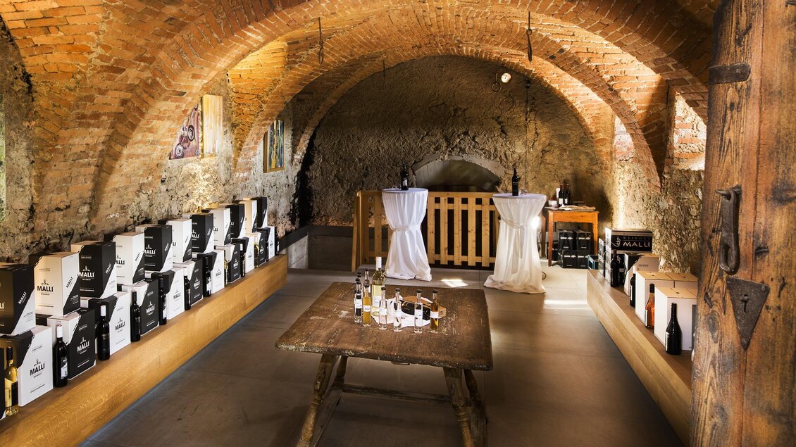 Ein stilvoller Weinkeller mit gewölbter Decke und Holzregalen. Auf einem Tisch sind Weinflaschen und Verkostungsutensilien zu sehen. | © Weingut Malli