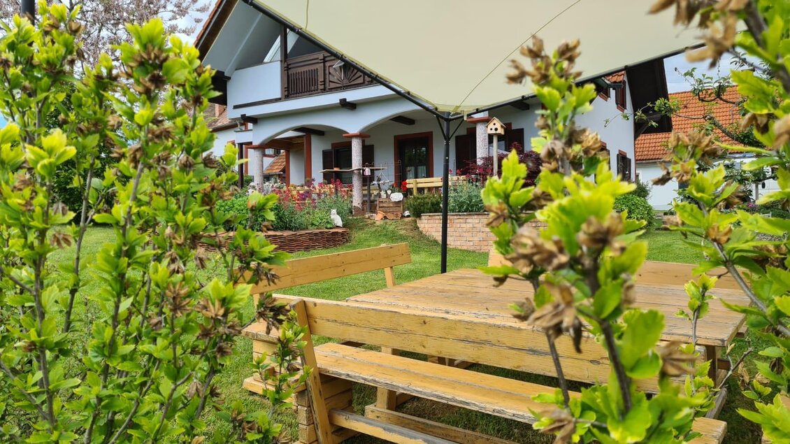 Ein gemütlicher Garten mit einem Holztisch und Bänken, umgeben von grünen Pflanzen. Im Hintergrund sieht man ein schönes Haus mit einem Balkon. | © Gerhard Maurer
