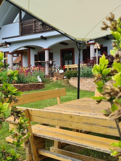 A cozy garden with a wooden table and benches, surrounded by green plants. In the background, there is a beautiful house with a balcony. | © Gerhard Maurer