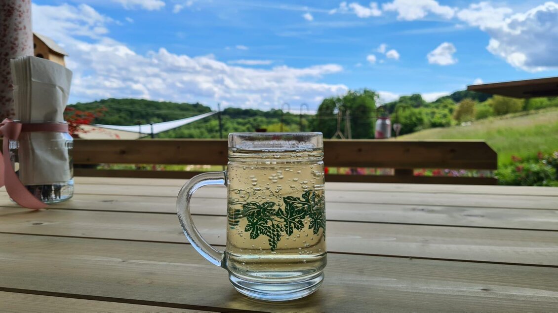 Ein Glas mit einem sprudelnden Getränk steht auf einem Holztisch. Im Hintergrund sind grüne Hügel und ein blauer Himmel mit vielen Wolken zu sehen. | © Gerhard Maurer