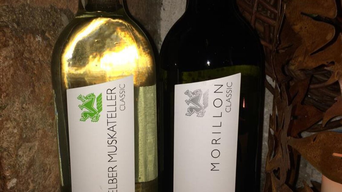 Zwei Flaschen Wein stehen auf einem rustikalen Untergrund. Die Flasche links ist ein weißer Gelber Muskateller und die rechte Flasche ist ein roter Morillon. | © Buschenschank Musterstubn l Nina & Armin Polz-Kiefer