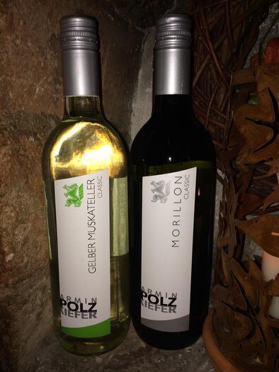 Zwei Flaschen Wein stehen auf einem rustikalen Untergrund. Die Flasche links ist ein weißer Gelber Muskateller und die rechte Flasche ist ein roter Morillon. | © Buschenschank Musterstubn l Nina & Armin Polz-Kiefer