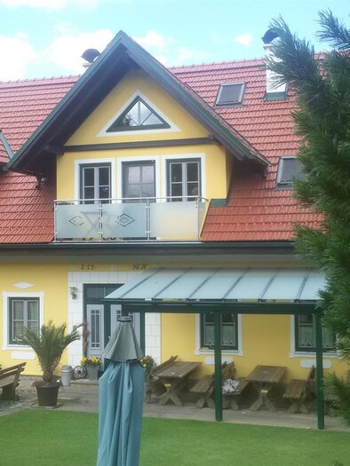 A beautiful house with a yellow facade and a red roof. In front of the house, there is a terrace and outdoor seating. | © Ofnerpeter
