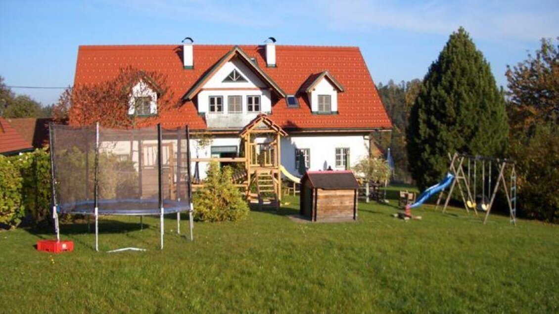 Ein schönes Einfamilienhaus mit rotem Dach in einem großen Garten. Im Garten befinden sich ein Trampolin, eine Rutsche und eine Schaukel. | © Buschenschank Ofnerpeter