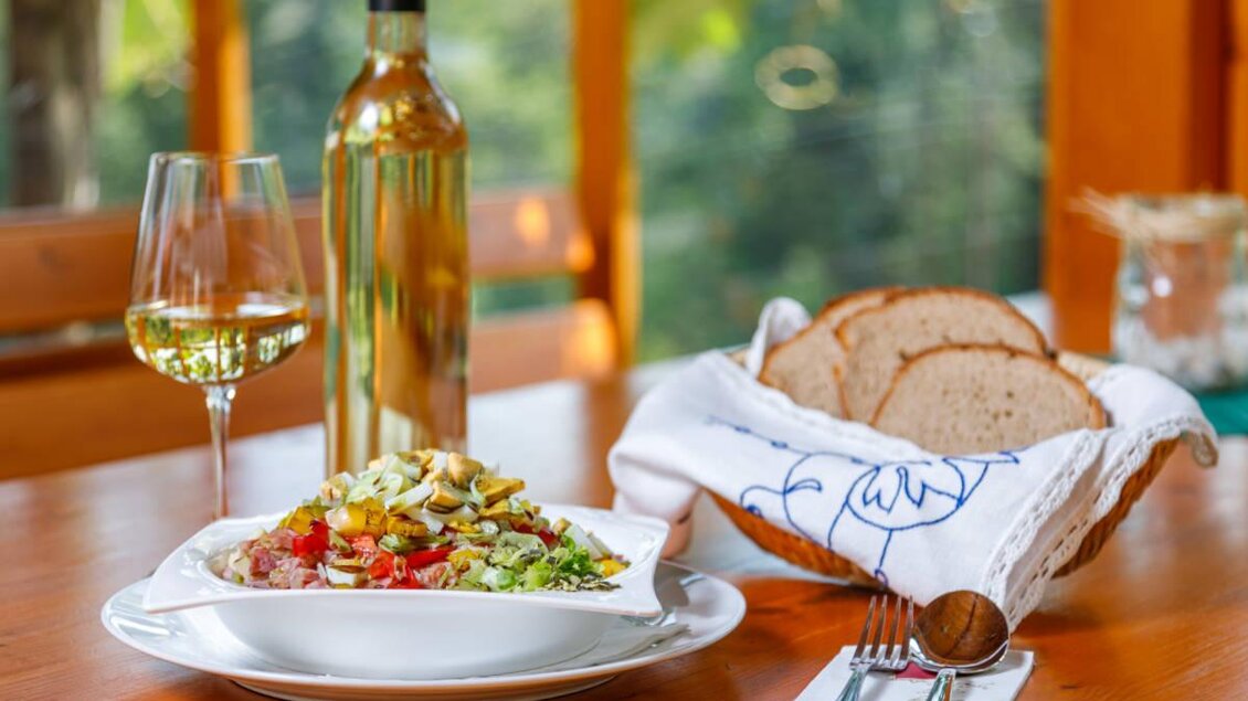 Ein Teller mit frischem Salat, kombiniert mit Gemüse und Nüssen, steht auf einem Holztisch. Daneben befinden sich ein Glas Weißwein und ein Korb mit Brot. | © Weinbau Pühringer