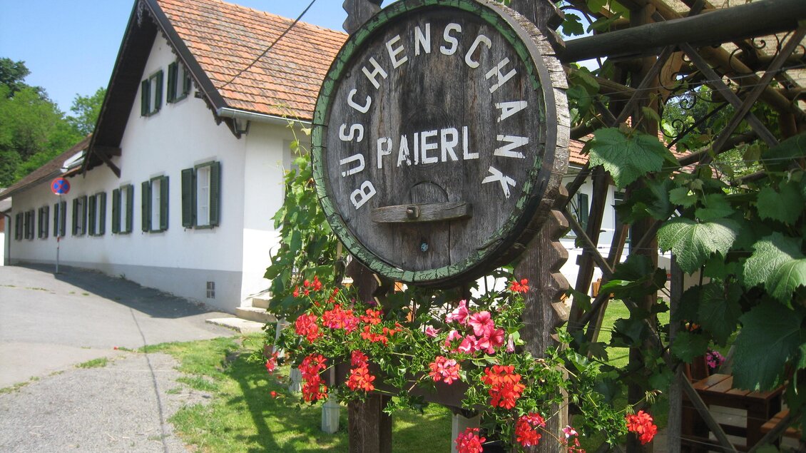 Ein rustikales Schild mit der Aufschrift „Buschenschank Paierl“ hängt an einer Holzkonstruktion. Im Hintergrund sieht man ein gemütliches Gebäude mit grünen Fensterläden. | © Buschenschank Paierl