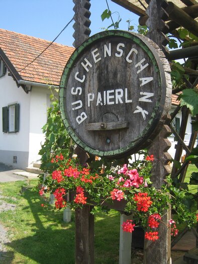 A rustic sign reading "Buschenschank Paierl" hangs from a wooden structure. In the background, you can see a cozy building with green shutters. | © Buschenschank Paierl