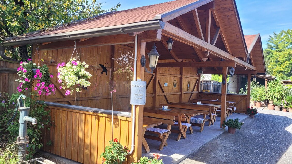 Ein gemütlicher Holzpavillon mit Tisch und Bänken im Freien. Blumenampeln verleihen dem Ort eine einladende Atmosphäre. | © TV Südsteiermark - Irene Löschnig