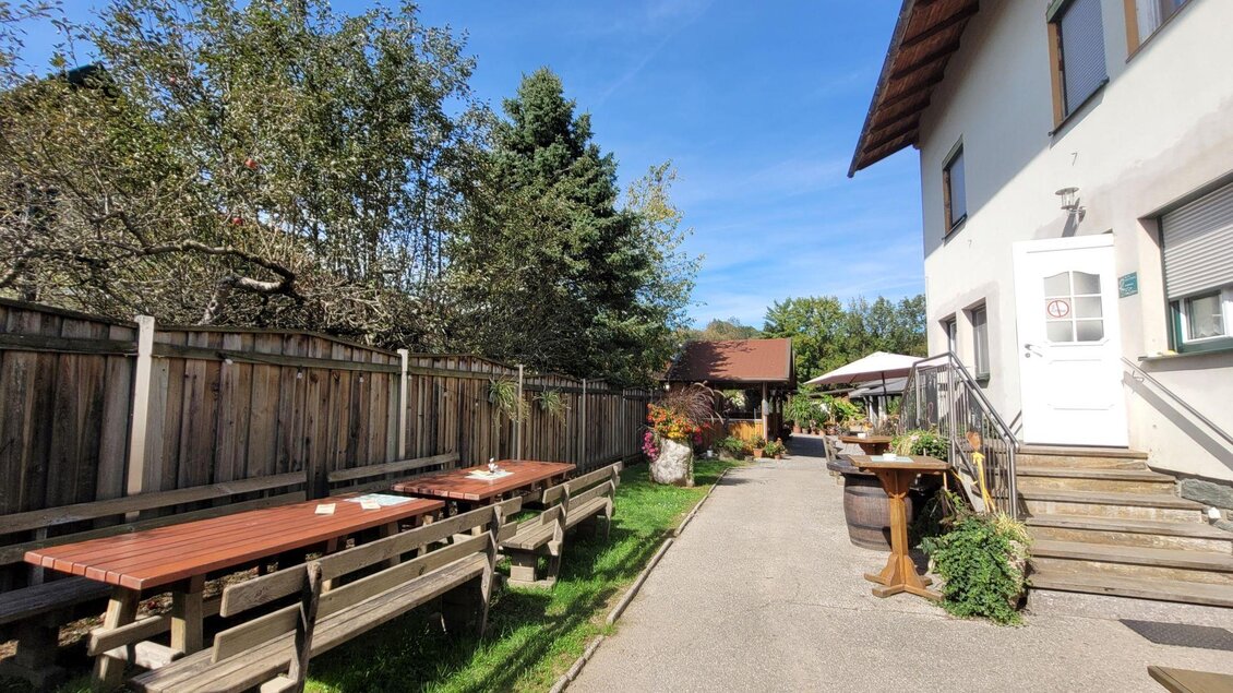 Ein einladender Garten mit Holztischen und Bänken. Im Hintergrund ein helles Gebäude und ein blauer Himmel. | © TV Südsteiermark - Irene Löschnig