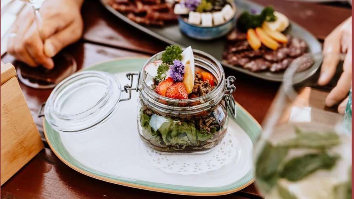 Ein dekoratives Glas mit frischen Salatzutaten und essbaren Blüten steht auf einem Teller. Im Hintergrund sind verschiedene appetitliche Gerichte zu sehen. | © Familie Repolusk | Weingut Repolusk