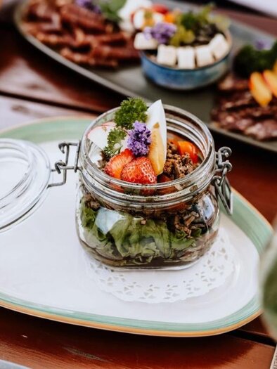 A decorative glass with fresh salad ingredients and edible flowers is placed on a plate. In the background, various appetizing dishes can be seen. | © Familie Repolusk | Weingut Repolusk