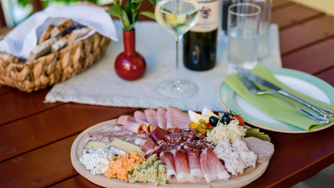 Eine stilvolle Platte mit verschiedenen Wurst- und Käsesorten, garniert mit buntem Gemüse. Im Hintergrund stehen Weingläser und eine Flasche Wein auf einem Holztisch. | © Familie Repolusk | Weingut Repolusk